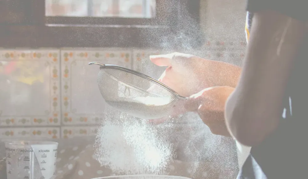Händer som siktar mjöl genom en finmaskig sil över en skål i ett kök